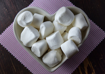 Fluffy white marshmallow in heart shapedd bowl on old wooden table with copy space close up.