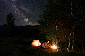 Four friends tourists having a rest by the campfire, sitting on logs during night camping among trees near the illuminated tent in the mountains under incredible beautiful starry sky with Milky way