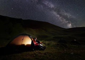 Fotobehang Man and woman having a rest at night camping in the Carpathians mountains near Dogyaska lake, sitting in tent, looking at the sky strewn with stars. In the tent lights on. Concept of active recreation © anatoliy_gleb