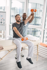 Weights training. Nice pleasant bearded man sitting on the medical couch and lifting his arm with a dumbbell