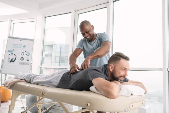 Relaxing atmosphere. Happy delighted man smiling while doing a back massage for his patient