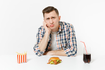 Handsome sad upset young man sitting at table with burger, french fries, cola in glass isolated on white background. Proper nutrition or American classic fast food. Advertising area with copy space.