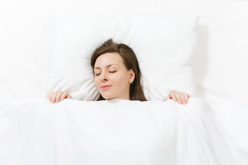 Obraz na płótnie Canvas Top view of head of happy brunette young woman lying in bed with white sheet, pillow, blanket. Sleeping pretty female spending time in room. Rest, relax good mood concept. Copy space for advertisement