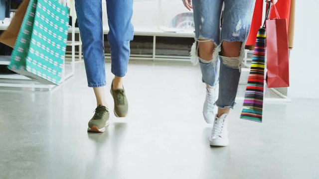 Low view of girls' legs walking slowly through spacious shop. Ladies are wearing jeans and modern trainers and carrying colourful paper bags.