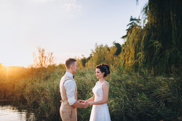 Lovely bride and groom joined hands and smiling at each other against the background of reeds and a lake. Newlyweds at sunset. Summer wedding.