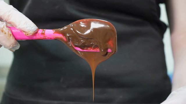 The brown mealted chocolate is dripping from the pink confectionary spoon. The pastry chief in black dickey is holding the tool by the hand in the white silicone glove. The candy paste is pouring down