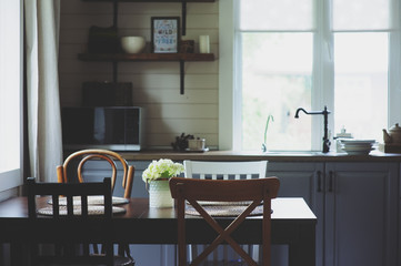 cozy summer morning at rustic country house kitchen. Wooden table with bouquet of fresh flowers, open shelving. Casual breakfast in grey and brown interior.