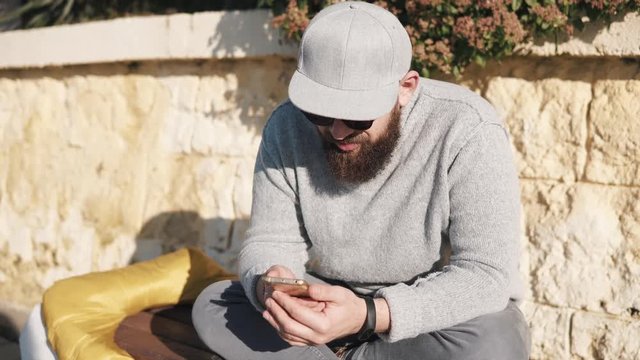 Grown man wearing grey sweater, jeans, cap and shandes sits on a bench outdoor under the sun. He is holding smartphone and chatting.