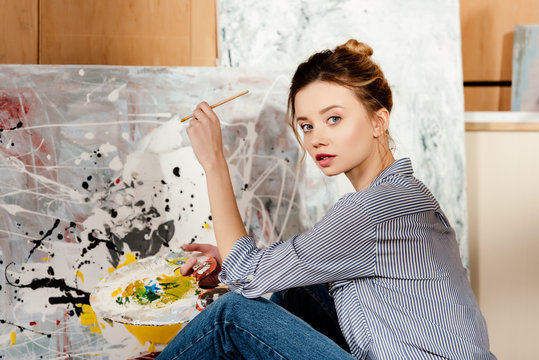 beautiful young artist holding palette and looking at camera in art studio