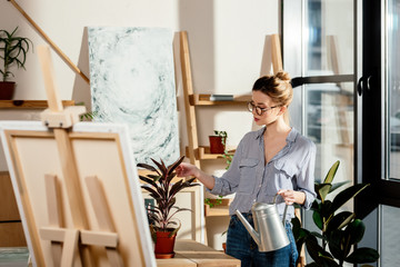 attractive stylish female artist in eyeglasses holding watering can and touching plant