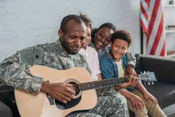 African American woman and children listening to father in camouflage clothes playing guitar