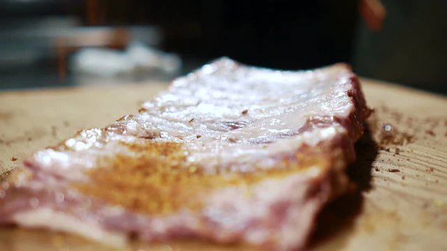 Young chef sprinkles spices on slices of meat in kitchen of restaurant. Male cook seasoned meat fillet with seasonings on table indoors. On wooden surface are delicious steaks, employee makes final