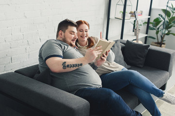 Obraz premium Smiling overweight boyfriend and girlfriend reading book on sofa at home