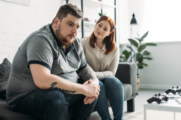 overweight girlfriend looking at sad boyfriend at home - obrazy, fototapety, plakaty