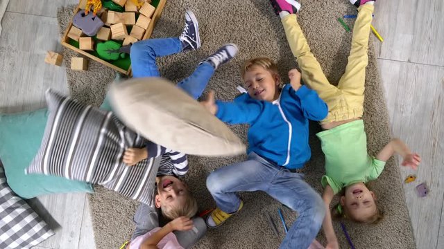 Group of four primary school friends lying on carpet on the floor and having fun with pillows that are falling down on them, top view