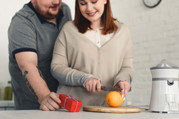 Cropped image of boyfriend presenting gift to overweight girlfriend while she cutting orange at kitchen