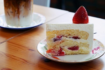 A strawberry shortcake topped with a large fresh strawberry placed in white plate and on wooden table with cafe environment.Victorian sponge cake with cream and strawberries.
