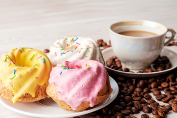 Three multi-colored cupcakes with icing and cup of coffee with milk on light table. Near it scattered coffee beans. Close-up view.