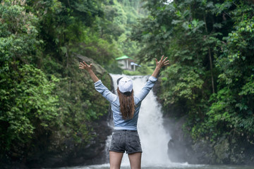 back view of woman with outstretched arms with Aling-Aling Waterfall on background, Bali, Indonesia