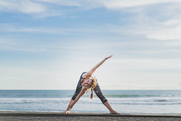 woman in Extended Triangle yoga pose with ocean and blue sky on background, Bali, Indonesia