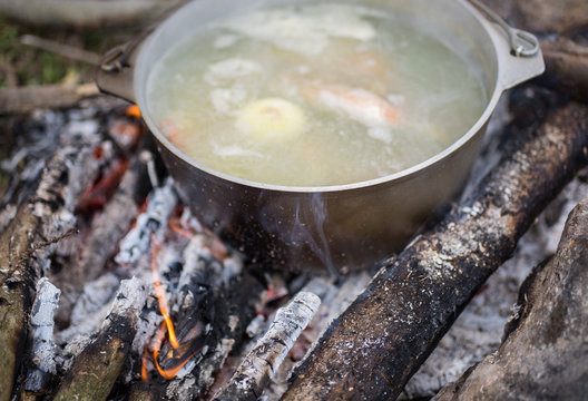 Old vintage pot in black soot is standing on the open fire in the wood. Natural summer holidays photo. Fish broth cooked in the forest. Vegetarian carrots and potatoes vegetable soup.