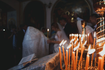 Marriage in orthodox church. Couple is walking in the cathedral with bright yellow candles on background. Priest in the long robe is marrying bride and groom couple.