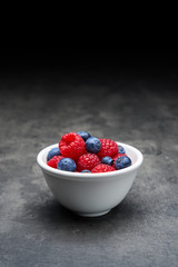 White ceramic bowl with berry fruit, blueberries and raspberries on dark concrete background. Close up with copy space at the top.