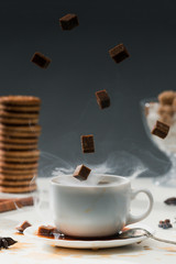 Steaming coffee cup with falling cane sugar cubes by cookies on table