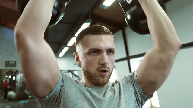 Portrait of male athlete, who is doing exercises with kettles in modern gym. Yound sportsman is holding weight equipment in his hands and comining squats and presses to work on the muscules of his
