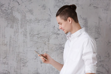 Side view of positive young man in casual clothes smiling and looking social network in smartphone on gray vintage wall background. Copyspace