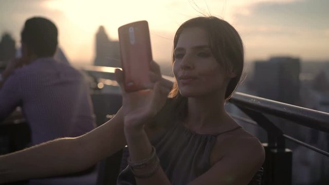 Young woman with drink taking selfie with cellphone in skybar during sunset
