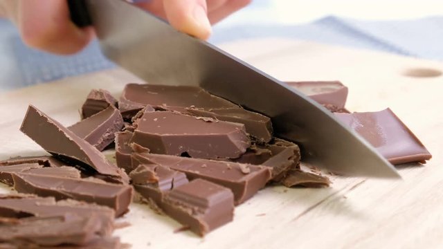 Cook hands cutting chocolate bar with a kitchen knife on cutting board.