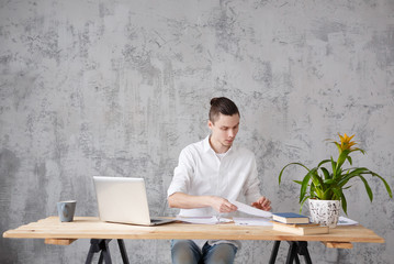 Cute young male writer while working on new scientific book sitting at the desk with laptop and books on the gray vintage background. Copyspace