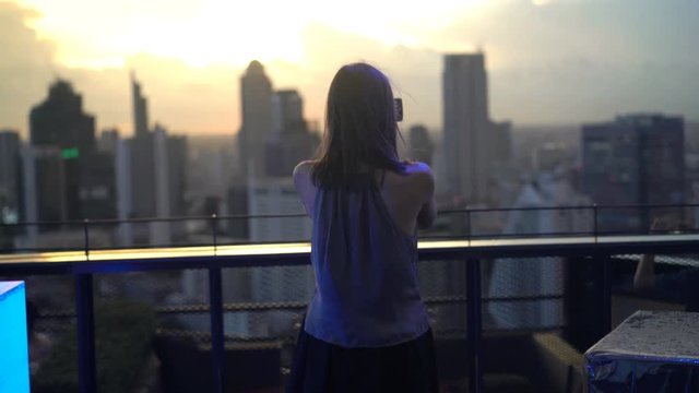 Young woman taking photo of cityscape with cellphone in skybar during sunset
