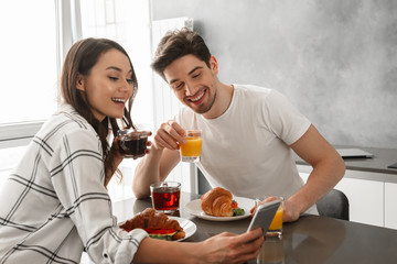 Portait of loving couple man and woman taking selfie on mobile phone, while having breakfast in kitchen