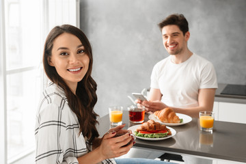 Portait of joyous man and woman 30s having breakfast or dinner in gray apartment, while sitting at table in kitchen