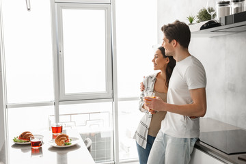 Picture of lovely couple smiling, and looking through big window, while having breakfast in modern apartment in morning