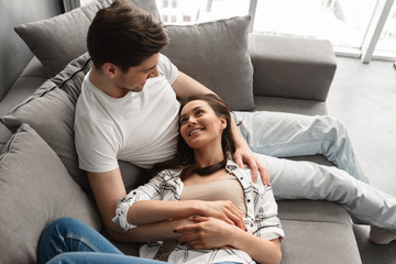 Photo of loving husband and wife in casual clothing resting in living room in modern flat, and lying on sofa together