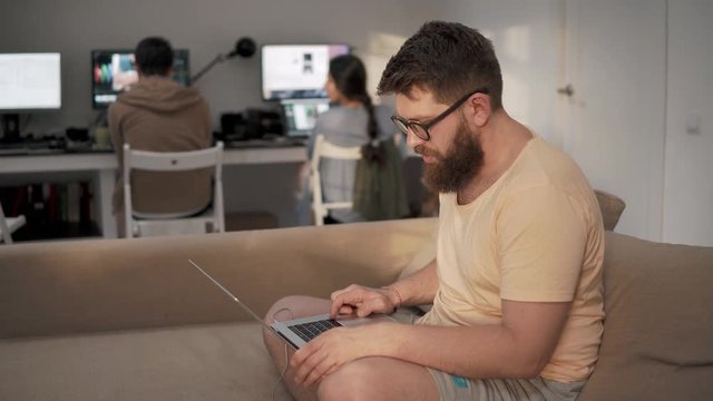 a young man with a beard on his face works in a friendly team, the person is dressed in homemade clothes and holds a laptop on his hands, he sits on the couch, behind him are computers for programming