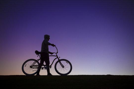Silhouette of a man in a hoodie standing with a cruiser bike in front of a purple sunset sky