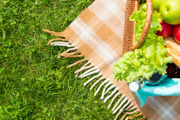 Green grass and straw basket with food and drinks on checkered beige tablecloth background for picnic, top view