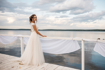 Fototapeta na wymiar Beautiful bride stands on the pier. Water and cloudy sky on the background