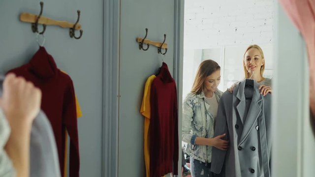Young lady in casual clothes is trying coat in fitting room and asking her friend for advice. Girl is coming closer, checking garment and approving it with smile.