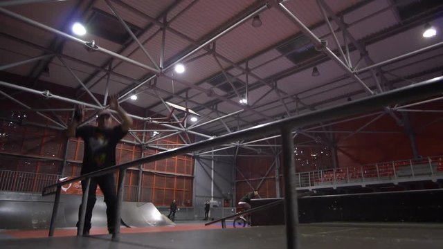 A man on rollers in a skate park performs a grind, in the background a person rides a bmx, slow motion