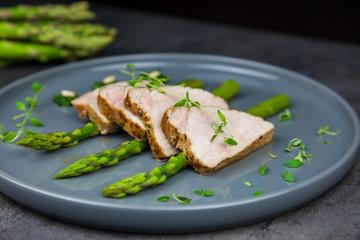 Slices of roasted pork tenderloin on asparagus with pesto from kale and thyme on the grey plate. A bunch of asparagus on the background