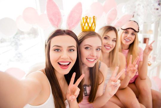 Attractive, charming, cheerful, pretty, stylish girls with crown, bunny ears, eye mask on heads shooting selfie on front camera showing v-sign, having theme party, dress code, sitting on bed