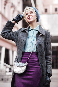 Outdoor portrait of yong beautiful happy smiling girl walking in street. Model wearing stylish pastel blue beret, blouse, violet suede skirt, gray coat, with small crossbody silvery bag