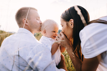 Walk beautiful young family in white clothes with a young son blond in mountainous areas with tall grass at sunset. Parents from both sides embracing son, hugging. family - this is happiness