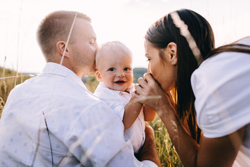 Walk beautiful young family in white clothes with a young son blond in mountainous areas with tall grass at sunset. Parents from both sides embracing son, hugging. family - this is happiness