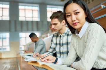 Portrait of two students sitting in row at desk in lecture hall of modern college, focus on young Asian woman looking at camera, copy space